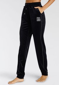 Schwarze Samtjogginghosen mit einem Kordelzugbund, seitlichen Taschen und dem Schriftzug "FEEL GOOD", der mit Strasssteinen entlang der Seitennaht verziert ist.
