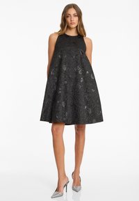 Femme portant une robe noire sans manches, arrivant aux genoux, avec un motif floral subtil et des chaussures à talons aiguilles argentées à l'arrière, se tenant sur un fond blanc.