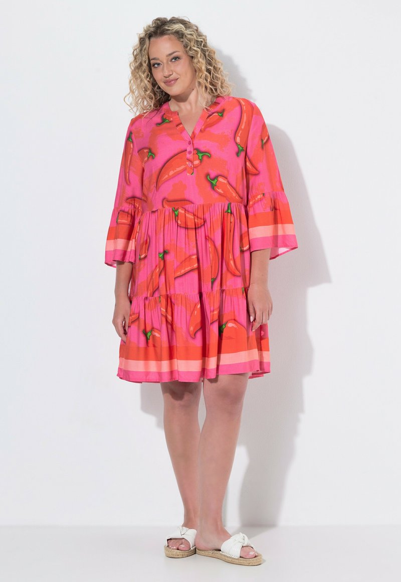 Woman with curly hair wearing a pink and orange chili pepper patterned dress and white sandals, standing against a plain background.