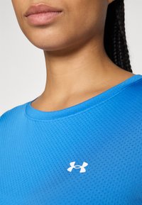 Kvinna klädd i en blå atletisk tröja med en vit Under Armour-logga, utsmyckad med små perforeringar för andningsförmåga.