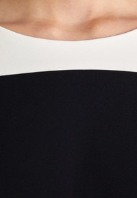 Schwarzes Kleid mit einem breiten weißen Ausschnitt, aus glattem Stoff gefertigt. Es hat ein eng anliegendes schwarzes Oberteil und einen strukturierten unteren Abschnitt.