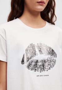 Hvit T-skjorte med korte ermer som har et grafisk design av overlappende tekst i grått, med teksten "Pepe Jeans London" tydelig vist.