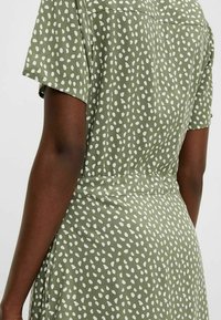 Robe en tissu vert avec un motif à pois blancs, à manches courtes et taille cintrée. La texture est lisse et légère.
