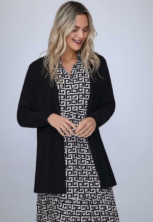 Cardigan nero sopra un vestito a fantasia in bianco e nero con chiusura a bottoni. Tessuto morbido, vestibilità rilassata e design aperto sul davanti.