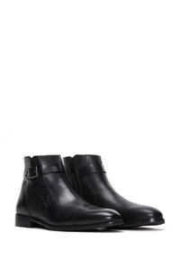 Botas tobilleras de cuero negro con cremallera lateral, correa decorativa con hebilla y paneles texturizados sobre un fondo blanco.