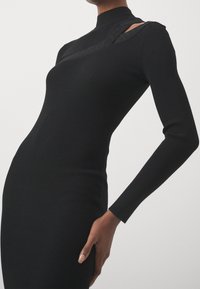 Robe noire côtelée avec col montant, manches longues et découpe à l'épaule. Le tissu est lisse avec une silhouette structurée.