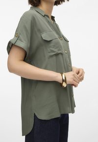 Camicia verde oliva a maniche corte con bottoni, dotata di due tasche sul petto e risvolti alle maniche, realizzata in tessuto leggero. Bracciale metallico dorato al polso.