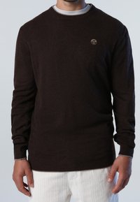 Pull en tricot marron avec un col rond, des manches longues et un logo discret sur la poitrine. Il présente une texture lisse et des poignets côtelés.