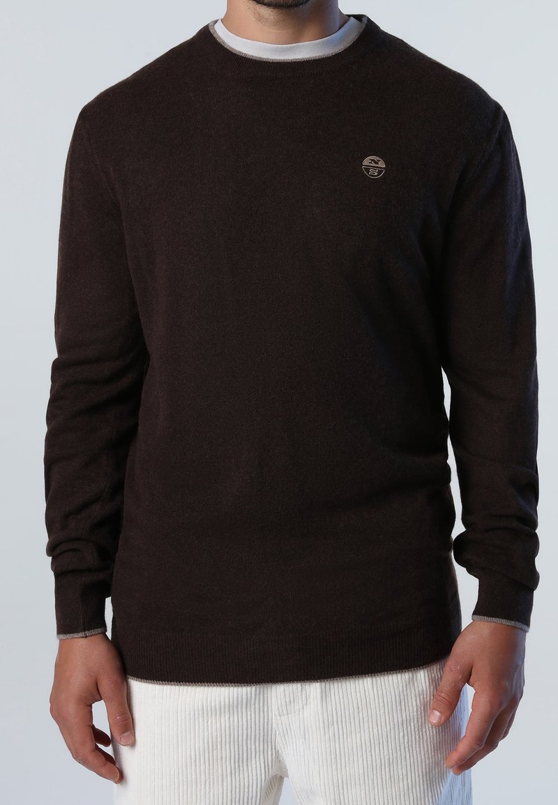 Pull en tricot marron avec un col rond, des manches longues et un logo discret sur la poitrine. Il présente une texture lisse et des poignets côtelés.