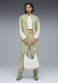 Lichtgroen twee-delig trainingspak met crèmekleurige accenten, bestaande uit een cropped jack en een broek met elastische tailleband. Voor wordt een witte schoudertas vastgehouden.