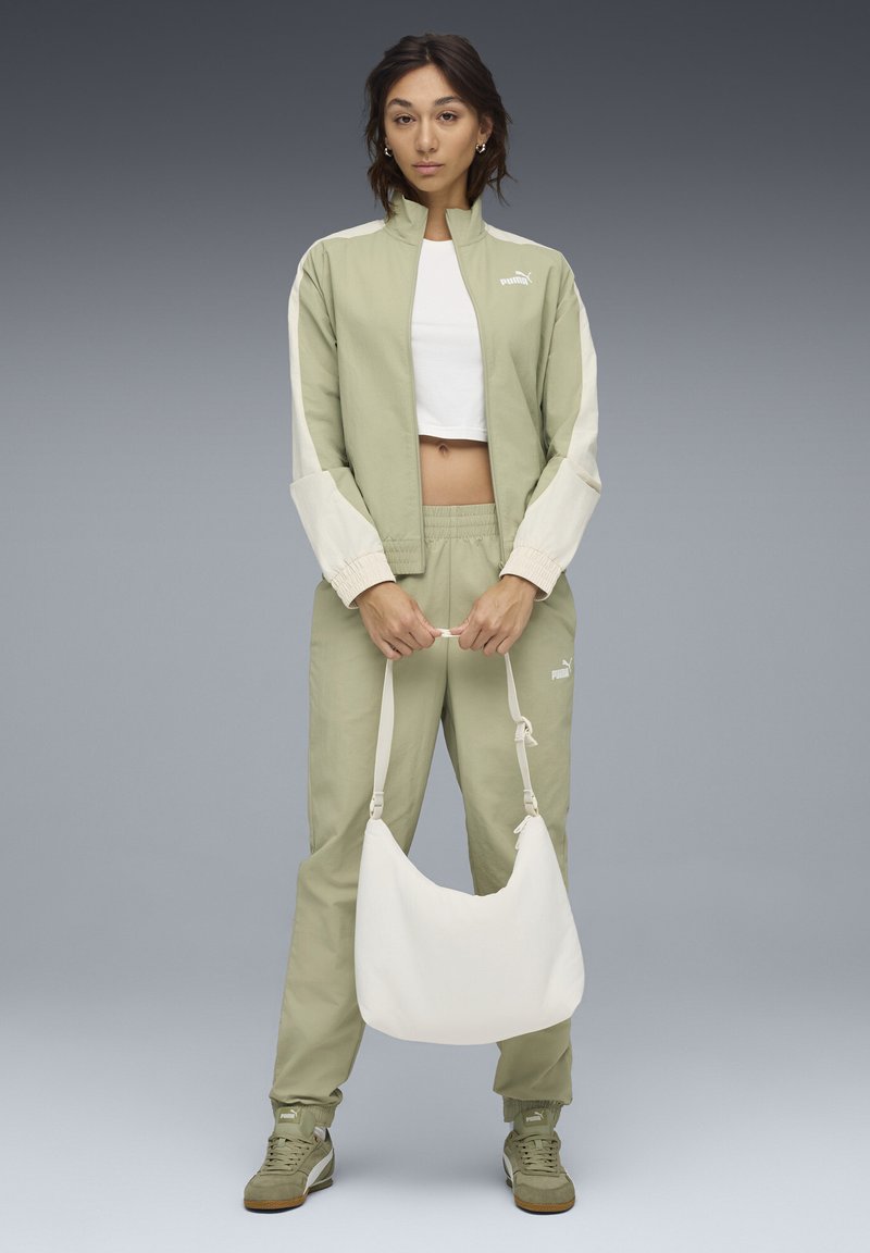 Lichtgroen twee-delig trainingspak met crèmekleurige accenten, bestaande uit een cropped jack en een broek met elastische tailleband. Voor wordt een witte schoudertas vastgehouden.