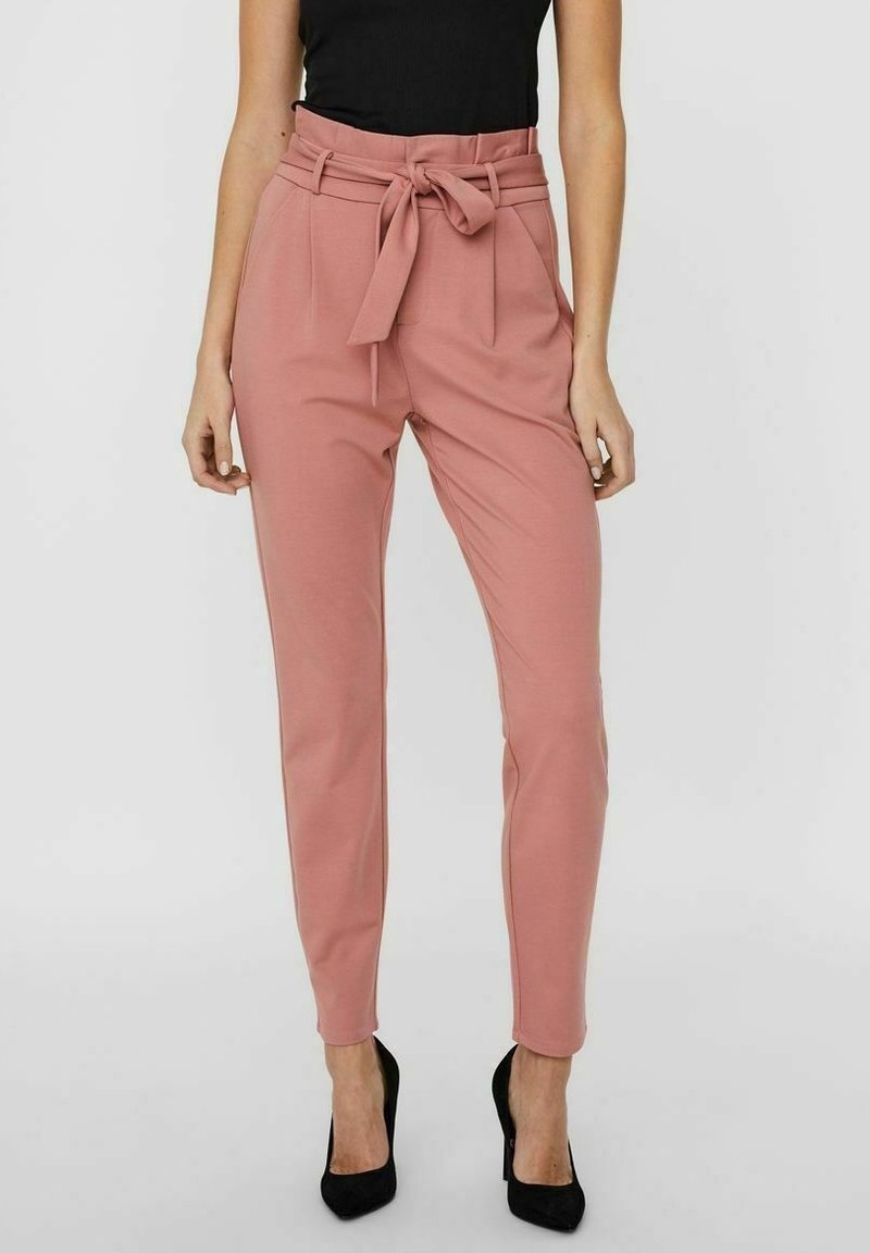 Pantalon rose sur mesure avec une taille haute et un détail de nœud. Tissu lisse, jambe fuselée, poches latérales, porté avec des talons hauts noirs.