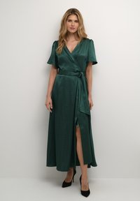 Rochie înfășurată din satin de culoare verde închis, cu mâneci scurte, talie legată și o fanta pe lateral, asortată cu pantofi cu toc înalt negri. Textură netedă.