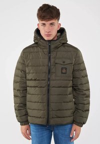 Giacca puffer verde oliva con cappuccio, chiusura a zip e tasca frontale con patch del logo. La cucitura orizzontale aggiunge texture.