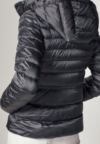 Sort pufferjakke med hætte, der har et kviltet design, skinnende tekstur og ribbede sømme. Bagfra ses en tætsiddende silhuet og søm.