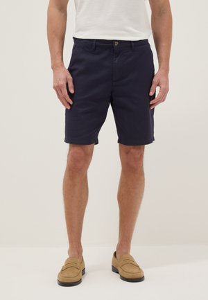 Homme portant un short bleu marine à taille plate, un t-shirt blanc et des mocassins beige, debout devant un fond clair uni.
