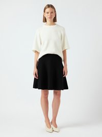 Femme debout portant un pull blanc à manches courtes, une jupe noire à longueur de genoux, des talons blancs et des boucles d'oreilles en argent contre un fond uni.