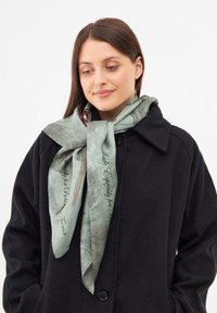 Lys grøn silkescarf med blomsterprint og tekst, elegant bundet om halsen, parret med en sort frakke for kontrast.