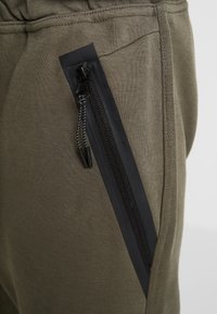 Olijfgroene sweatpants met een slanke zwarte ritszak aan de zijkant, gemaakt van zachte stof met een gladde textuur.