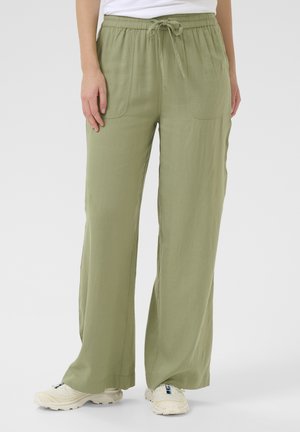 Person iført let olivengrønne bukser med vide ben, forlommer og snørelukning i taljen, kombineret med hvide sneakers og en hvid top.