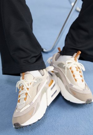 Person iført beige, hvide og tan sneakers med mesh- og ruskindsdetaljer, stående på lyseblåt gulv, iført sorte bukser.