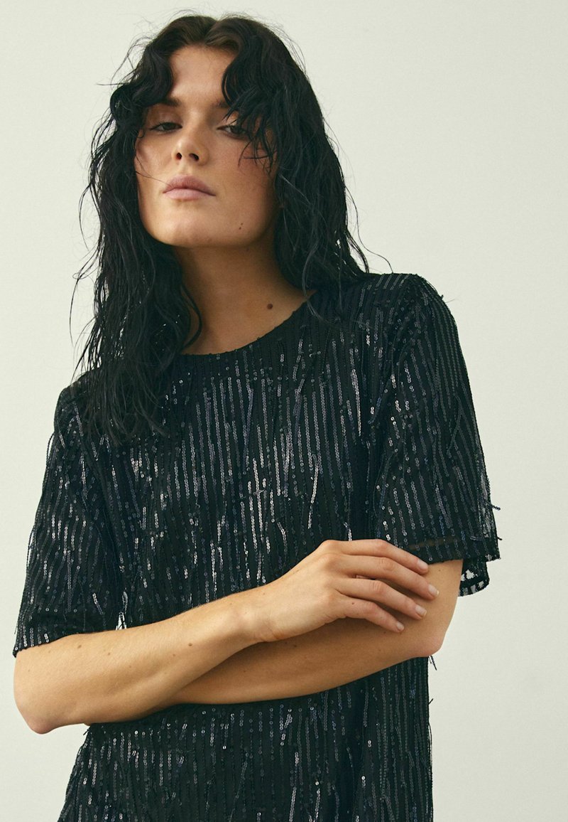 Schwarzes, mit Pailletten besetztes Oberteil mit kurzen Ärmeln, das ein lockeres Design und eine strukturierte Oberfläche aufweist, akzentuiert durch senkrechte Streifen aus schimmernden Pailletten.