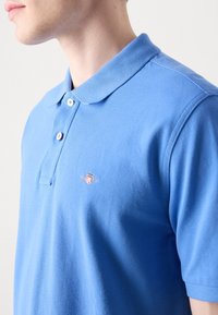 Blaues Poloshirt mit strukturiertem Finish, ausgestattet mit einem spitzen Kragen, zwei Knöpfen und einem kleinen gestickten Logo auf der linken Brust.