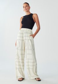 Zwarte geribbelde tanktop gecombineerd met ivoorkleurige broek met brede pijpen, versierd met zwarte geometrische lijnen en zakken, op een neutrale achtergrond.