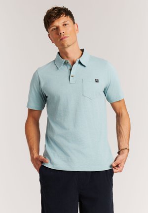 Man in lichtblauw poloshirt met zakje en donkere broek, staand met handen dicht bij de heupen tegen een effen achtergrond.