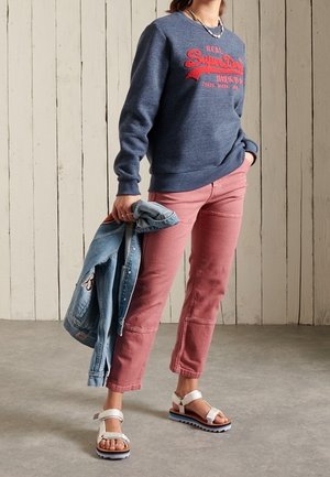 Sudadera de marina con gráfico rojo, pantalones rosa ajustados y sandalias blancas con suelas azules; chaqueta de mezclilla colgada sobre el brazo izquierdo.