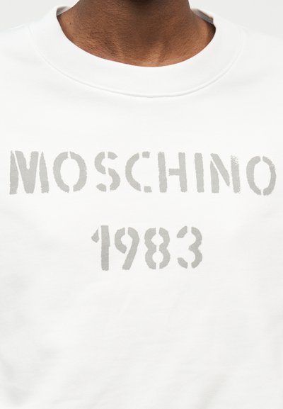 Λευκό φούτερ με στρογγυλή λαιμόκοψη και ξεθωριασμένο γκρι κείμενο "MOSCHINO 1983" στο στήθος, φορεμένο από άτομο με σκούρο χρώμα δέρματος.