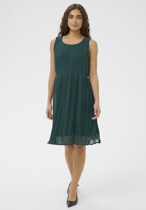 Kobieta z długimi kręconymi włosami, ubrana w bezrękawnikową, sięgającą kolan plisowaną sukienkę w ciemnozielonym kolorze oraz czarne, spiczaste buty, stojąca na jednolitym tle.