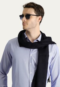 Sciarpa nera drappeggiata attorno al collo, realizzata in materiale texturizzato, abbinata a occhiali da sole grigi e a una camicia azzurro chiaro con bottoni.