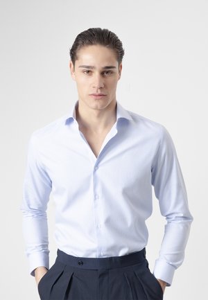 Joven con el pelo peinado hacia atrás, que lleva una camisa de botones azul claro y pantalones oscuros, de pie con las manos en los bolsillos frente a un fondo liso.