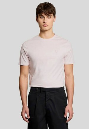 Joven con cabello corto castaño, vistiendo una camiseta de manga corta de color rosa claro y pantalones negros, de pie contra un fondo gris liso.