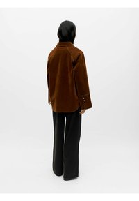 Brun fløyelskjorte med krage, lange ermer og knappedetaljer. Kombinert med svarte vide bukser. Ensfarget bakgrunn.