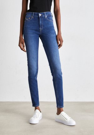 Person trägt eng geschnittene blaue Jeans und weiße Sneakers und steht auf einem hellgrauen Boden vor einer schlichten hellen Wand.