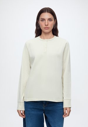 Femme aux longs cheveux bruns portant un t-shirt henley à manches longues beige clair et un jean bleu, regardant vers l'avant avec une expression neutre.