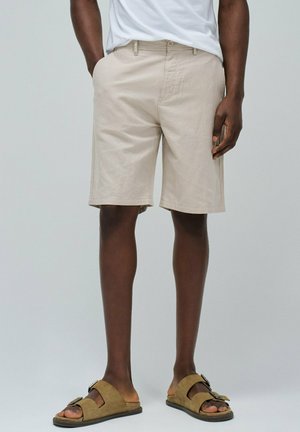 Pantalones cortos beige de corte recto con bolsillos laterales, combinados con sandalias marrones que tienen una correa y detalle de hebilla.