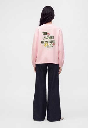 Vrouw met lang donker haar die een roze trui met bloemige tekst "The Flower Gardening Club" en wijde zwarte broek draagt, gezien van achteren.