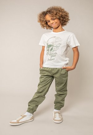 Enfant aux cheveux bouclés portant un t-shirt blanc avec un motif de crâne, un pantalon jogger vert et des baskets blanches, debout les mains dans les poches.