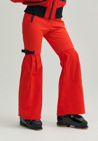 Pantaloni de schi roșii cu tiv evazat, având un detaliu cu cataramă la genunchi. Asortați cu cizme de schi negre și un design de țesătură texturată.