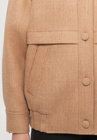 Világosbarna gyapjú dzseki, mely texturált anyagból készült, gombos záródással és két elülső zsebbels rendelkezik. A dizájnba beletartozik a hajtott gallér és a varrott varrások.