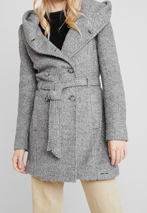 Žena nosící šedý strukturovaný vlněný kabát s páskem přes černý top a béžové kalhoty, stojící před jednoduchým šedým pozadím.