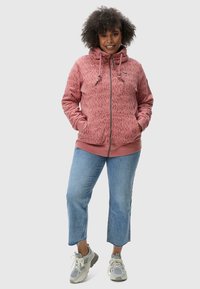 Felpa con zip rossa a fantasia, con collo alto e lacci, abbinata a jeans blu cropped e sneakers grigie.
