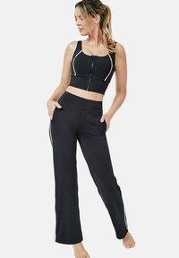 Zwarte sportieve crop top met rits en gouden accenten, gedragen met zwarte wijde broek met zijstrepen en zakken.