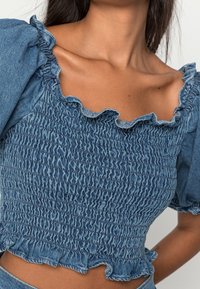 Haut en denim avec un design smocké, dotée d'un col froncé et de manches bouffantes, dans une teinte bleu moyen avec une finition texturée.