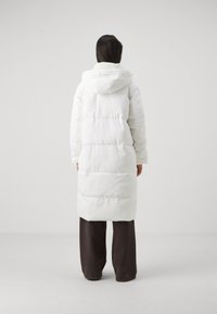 Manteau rembourré long blanc avec capuche, doté d'un design matelassé et d'une doublure en peluche, atteignant le mi-mollet, associé à un pantalon ample marron foncé.