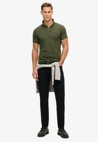 Polo verde oliva, de manga corta, con un pequeño logotipo. Pantalones negros con un cinturón, suéter gris claro atado a la cintura, zapatos negros.