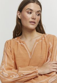 Ferskenfarvet linned blouse med lange, samlede ærmer, V-hals og hvide broderede mønstre langs halsudskæringen og ærmerne.